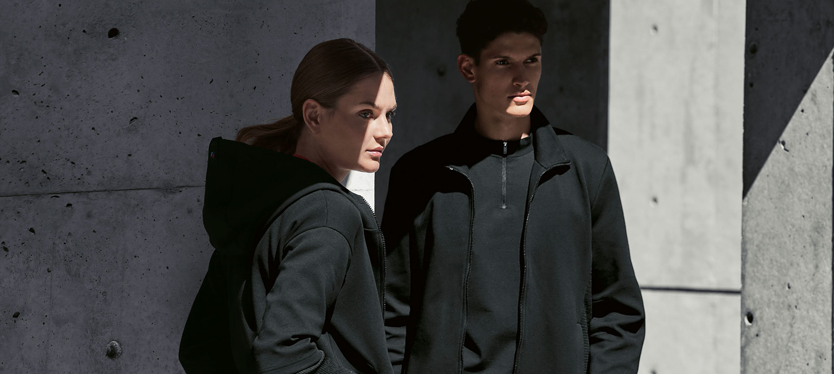 A couple ist standing in the shadow. A woman is presenting the BMW M Sweat Jacket for ladies and a man is wearing the BMW M Sweat Jacket for men.